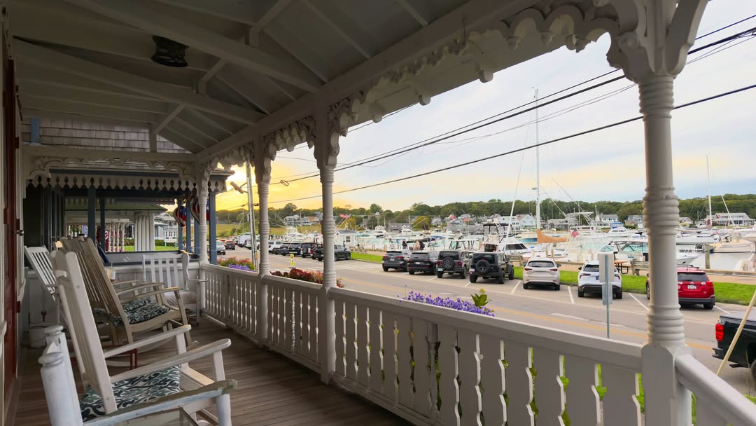 A gingerbread-trimmed porch with classic rocking chairs looking out over Oak Bluffs Harbor, sailboat...