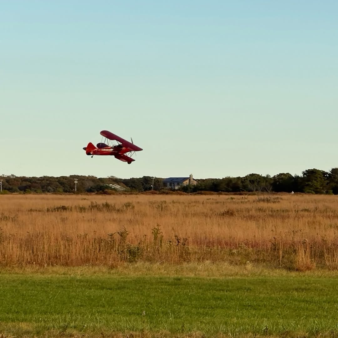 A red biplane low over Katama Airfield in Edgartown, with the field gone gold for late October. This...